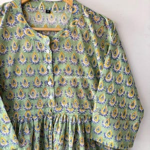 May include: A green cotton tunic with a floral print featuring yellow and blue accents. The tunic has a button-up front and a gathered waist.
