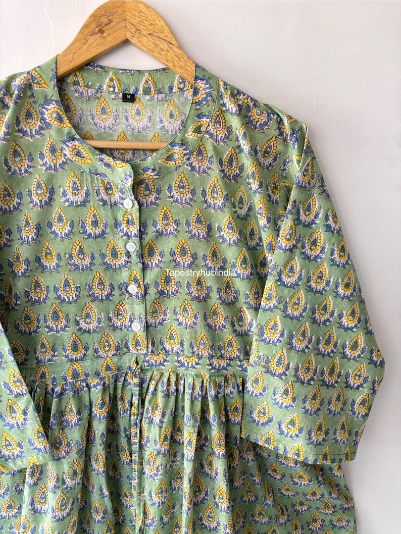 May include: A green cotton tunic with a floral print featuring yellow and blue accents. The tunic has a button-up front and a gathered waist.