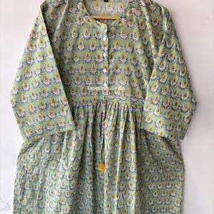 May include: A green cotton tunic with a yellow and blue floral print. The tunic has a button-up front and a gathered waist. The text "Tapestryhubindia" is visible on the front of the tunic.