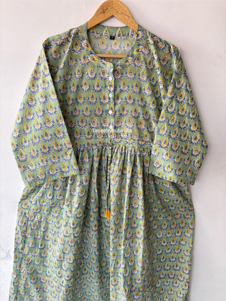 May include: A green cotton tunic with a yellow and blue floral print. The tunic has a button-up front and a gathered waist. The text "Tapestryhubindia" is visible on the front of the tunic.