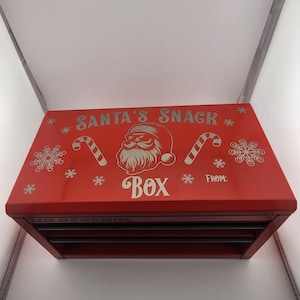 May include: A red metal box with the words "SANTA'S SNACK BOX" and a Santa Claus graphic. The box has candy cane and snowflake designs. The bottom of the box has multiple drawers.