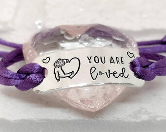 May include: Purple cord bracelet with a silver charm that says "You are loved" and has a small drawing of a sloth holding a heart.