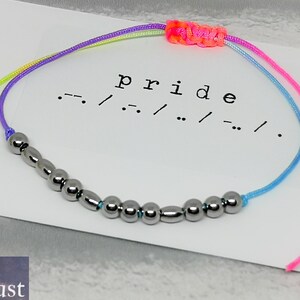 May include: A rainbow colored string bracelet with silver beads. The bracelet is tied with a knot and has the word "pride" written on a white card with the word "pride" spelled out in braille.