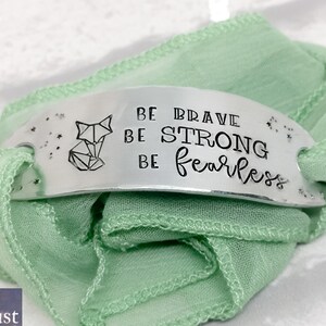 May include: A silver bracelet with a green ribbon. The bracelet has a geometric fox design and the words "Be Brave Be Strong Be Fearless".