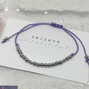 May include: A purple string bracelet with silver beads that spells out "believe" in braille. The bracelet is on a white card with the word "believe" written in black.