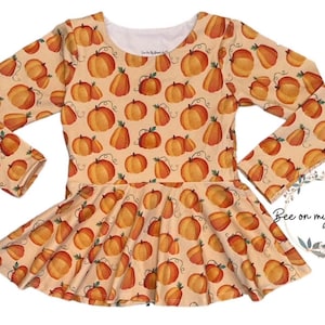 May include: A long-sleeved dress featuring a pumpkin pattern. The dress has a light tan background with orange pumpkins and green vines. The dress has a round neckline and a flared skirt. The text "Bee on my Bonnet Designs" is visible.