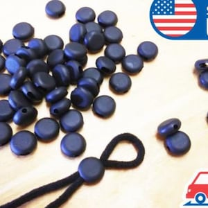 May include: A close-up of a group of black plastic cord ends. The cord ends are round and have a small hole in the centre. There are 40 cord ends in total. The cord ends are on a wooden surface. Ships from USA.