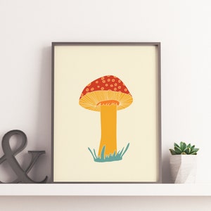 May include: A framed art print with a mushroom illustration. The mushroom has a red cap with white spots, a yellow underside, and an orange stem. Green grass is at the base of the stem, set against a cream background. The print is displayed on a shelf.