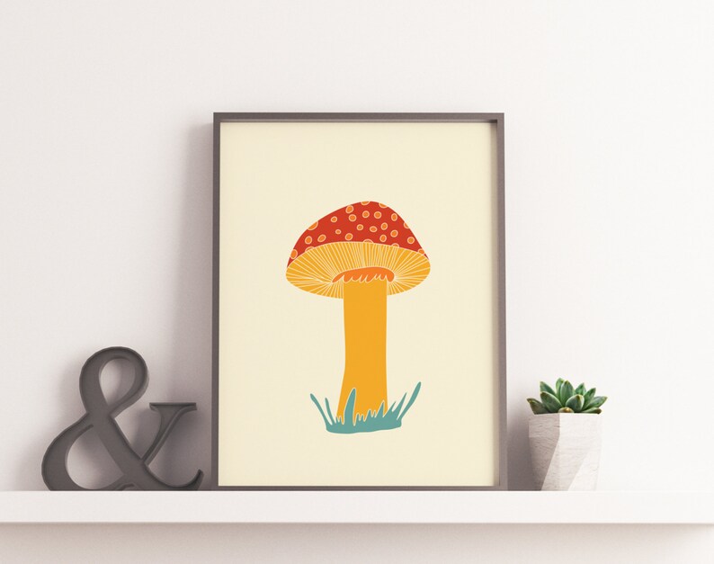 May include: A framed art print with a mushroom illustration. The mushroom has a red cap with white spots, a yellow underside, and an orange stem. Green grass is at the base of the stem, set against a cream background. The print is displayed on a shelf.
