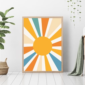 May include: A framed print of a stylized sun with orange, yellow, white, and blue rays. The sun is in the center of the print and is surrounded by rays that radiate outwards. The print is framed in a light wood frame.