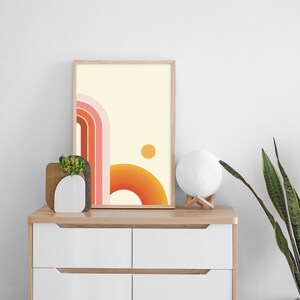 Pu&ograve; includere: Una stampa incorniciata con un design astratto in stile retr&ograve; che presenta una serie di linee curve in tonalit&agrave; di rosa, arancio e giallo. Il design ricorda un tramonto o un'alba con un piccolo cerchio nell'angolo in alto a destra.