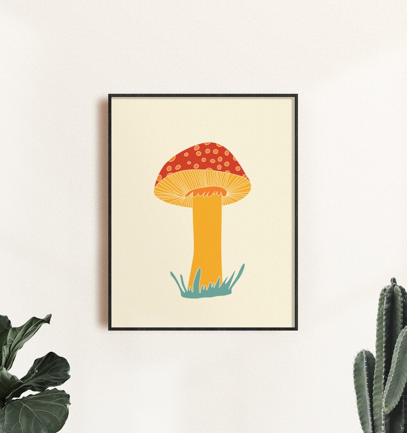 May include: A framed art print depicting a stylized mushroom illustration. The mushroom has a red cap with white spots, a yellow stem, and is set against a cream background. The artwork is framed in a black frame.