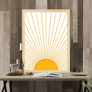 May include: A framed print of a stylized sun with yellow rays on a white background. The print is on a wooden table with a book, plants, and a candle.