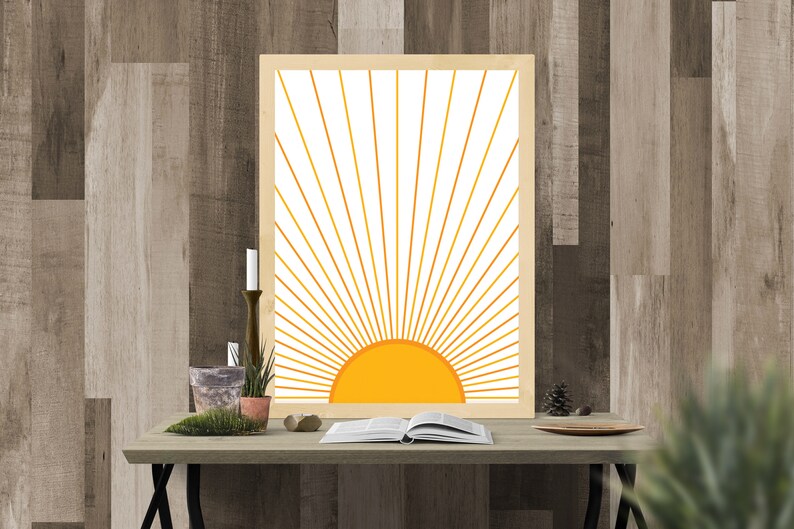 May include: A framed print of a stylized sun with yellow rays on a white background. The print is on a wooden table with a book, plants, and a candle.