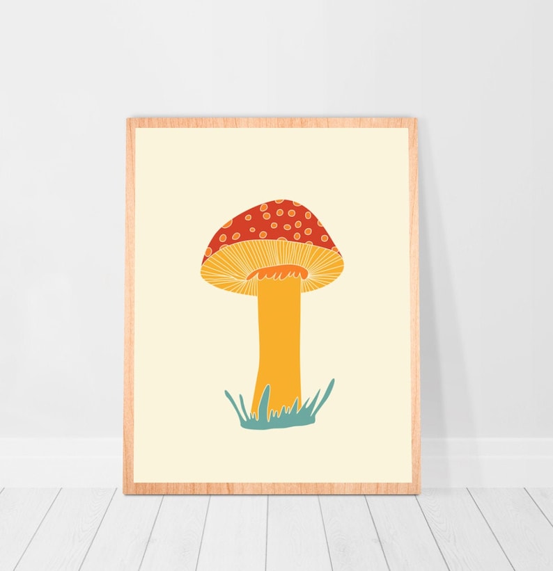 May include: A framed art print depicting a stylised mushroom illustration. The mushroom has a red cap with white spots, a yellow underside, and an orange stem. The artwork is set against a cream background, with blue-green grass at the base.