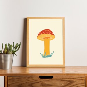 May include: A framed art print showing a stylised mushroom illustration. The mushroom has a red cap with white spots, a yellow underside, and an orange stem. The background is a light yellow colour.