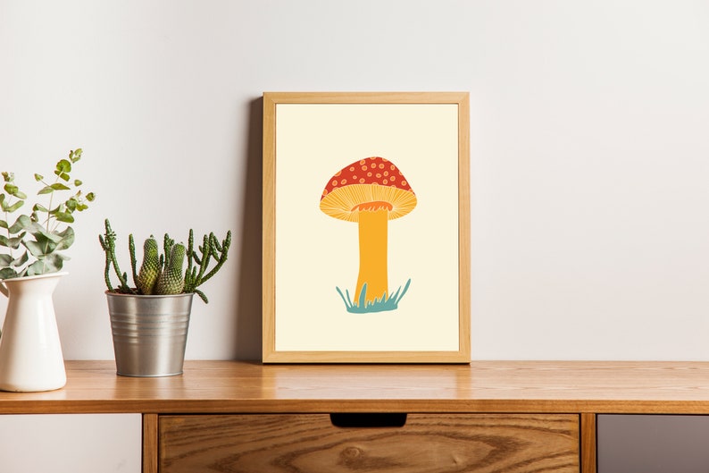 May include: A framed art print showing a stylised mushroom illustration. The mushroom has a red cap with white spots, a yellow underside, and an orange stem. The background is a light yellow colour.