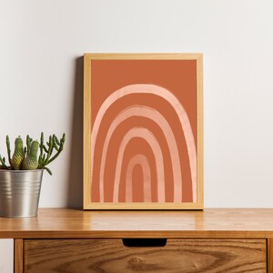 May include: A framed art print featuring a minimalist rainbow design in shades of peach and terracotta. The artwork is displayed on a wooden surface, alongside a potted cactus and a white vase with greenery, creating a warm, inviting aesthetic.