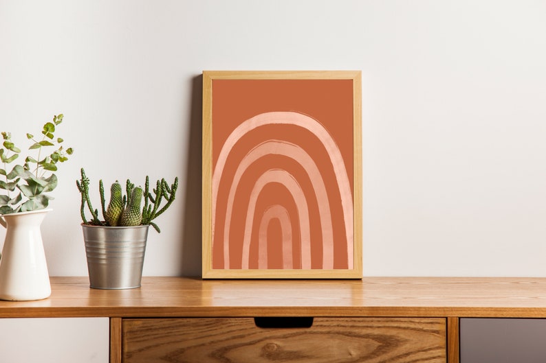 May include: A framed art print featuring a minimalist rainbow design in shades of peach and terracotta. The artwork is displayed on a wooden surface, alongside a potted cactus and a white vase with greenery, creating a warm, inviting aesthetic.