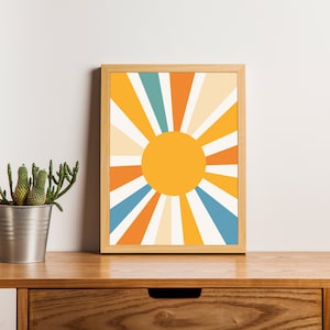 May include: A framed print of a stylized sun with orange, yellow, white, and blue rays. The print is in a wooden frame and is sitting on a wooden shelf with a potted plant.