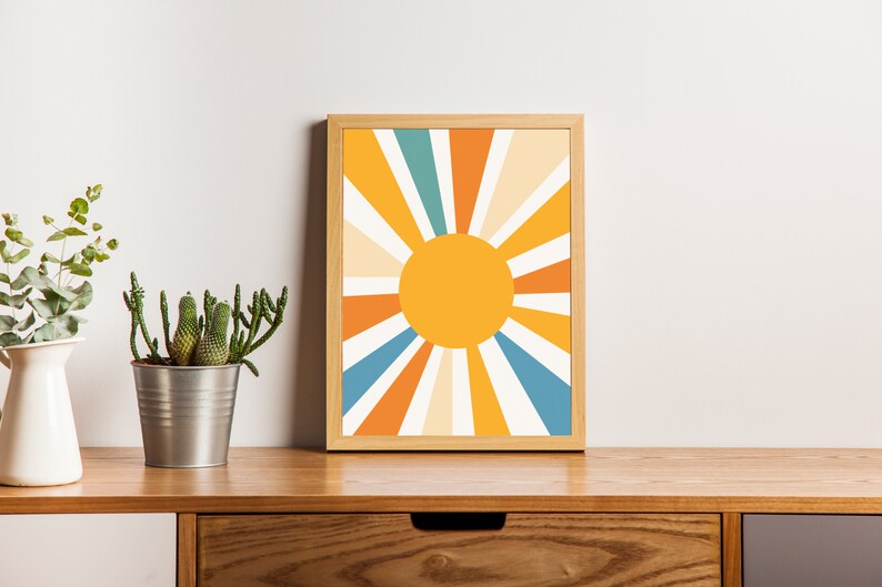 May include: A framed print of a stylized sun with orange, yellow, white, and blue rays. The print is in a wooden frame and is sitting on a wooden shelf with a potted plant.