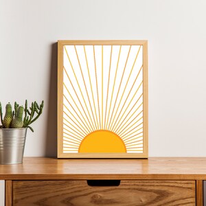 May include: A framed print of a stylized sun with yellow and orange rays on a white background. The frame is light wood.