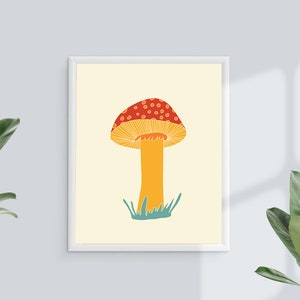 May include: A framed art print showing a stylized illustration of a mushroom. The mushroom has a red cap with white spots, a yellow underside, and a yellow stem. Green grass is at the base of the stem. The background is a light cream colour.
