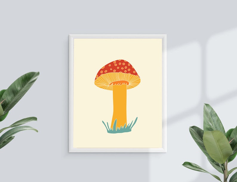 May include: A framed art print showing a stylized illustration of a mushroom. The mushroom has a red cap with white spots, a yellow underside, and a yellow stem. Green grass is at the base of the stem. The background is a light cream colour.