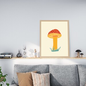 May include: A framed art print depicting a stylised mushroom illustration. The mushroom has a red cap with white spots, a yellow underside, and an orange stem. Green grass is at the base. The print is in a light brown frame.