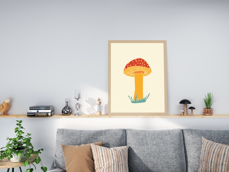 May include: A framed art print depicting a stylised mushroom illustration. The mushroom has a red cap with white spots, a yellow underside, and an orange stem. Green grass is at the base. The print is in a light brown frame.