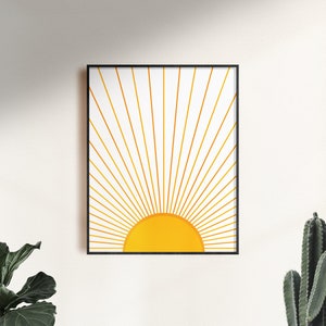 May include: A framed print of a minimalist sun design with gold rays and a yellow sun on a white background.