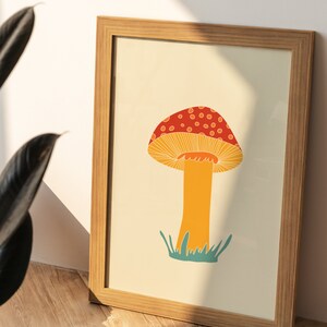 May include: A framed art print featuring a colourful mushroom illustration. The mushroom has a red cap with white spots, an orange stem, and is set against a cream background. The frame is made of light-coloured wood.
