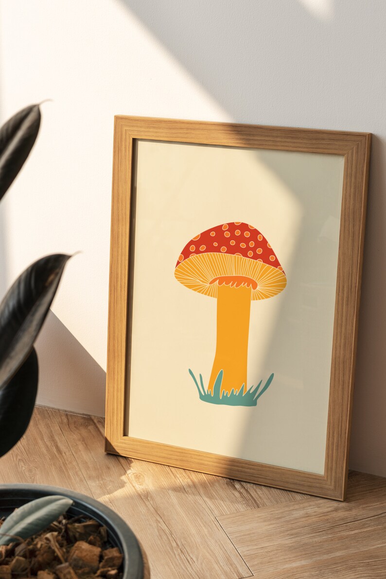 May include: A framed art print featuring a colourful mushroom illustration. The mushroom has a red cap with white spots, an orange stem, and is set against a cream background. The frame is made of light-coloured wood.