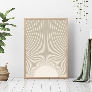 Puede incluir: Una impresi&oacute;n enmarcada de un dise&ntilde;o de sol beige con un sol blanco en el centro. La impresi&oacute;n est&aacute; colgada en una pared blanca con una planta verde en una cesta de mimbre y un sof&aacute; gris con una manta de tiro verde en primer plano.