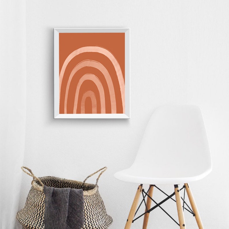 May include: A framed abstract art print featuring a rainbow design in shades of orange and peach. The artwork is displayed in a white frame. The image is set against a white wall, with a woven basket and a white chair in the foreground.