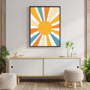 May include: A framed print of a stylized sun with orange, yellow, blue, and white rays. The print is hanging on a white wall above a light wood cabinet with white doors.