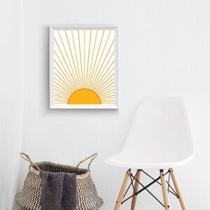May include: A white framed print of a stylized yellow sun with golden rays radiating outward. The print is hanging on a white wall above a white chair.