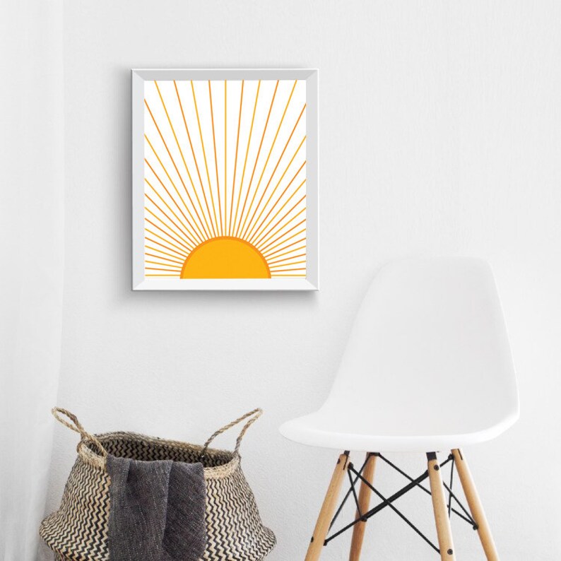 May include: A white framed print of a stylized yellow sun with golden rays radiating outward. The print is hanging on a white wall above a white chair.