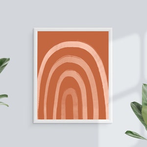 May include: A framed art print featuring a stylized rainbow design. The artwork showcases a series of curved lines in varying shades of orange and peach against a solid orange background. The print is encased in a white frame.