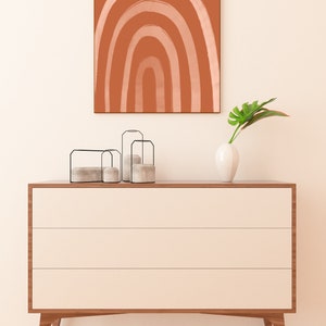 May include: A modern, three-drawer dresser with a light wood finish and cream-colored drawers. Above, a framed abstract art piece with a terracotta background and a white rainbow design. Decorative candles and a white vase with a green leaf complete the scene.