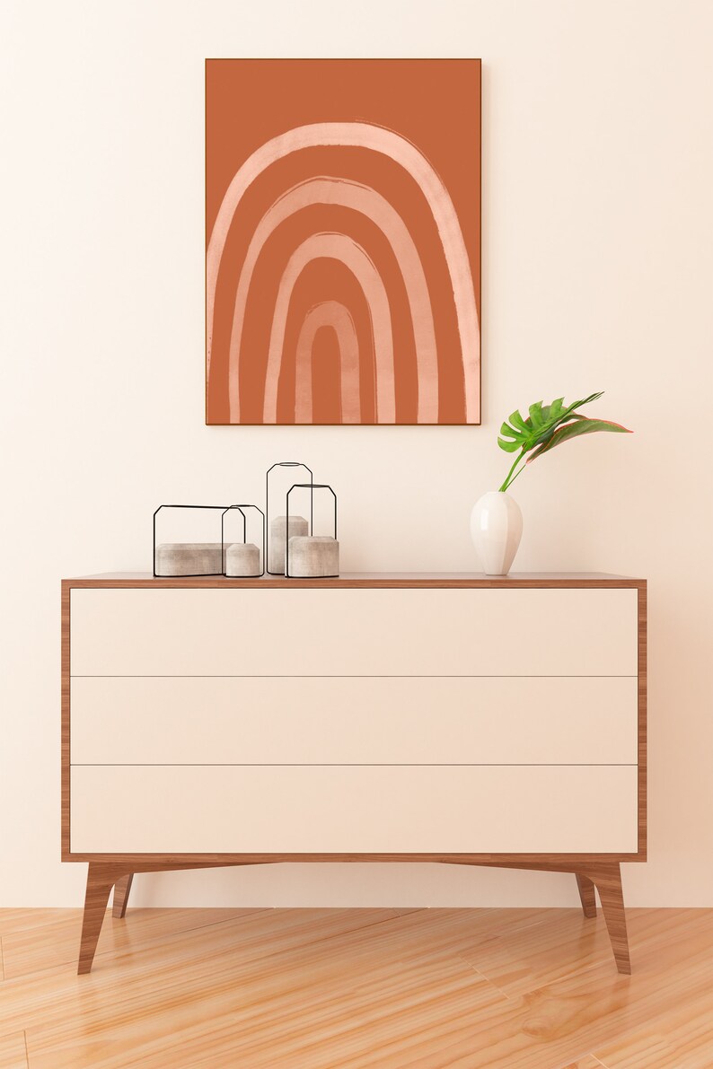 May include: A modern, three-drawer dresser with a light wood finish and cream-colored drawers. Above, a framed abstract art piece with a terracotta background and a white rainbow design. Decorative candles and a white vase with a green leaf complete the scene.