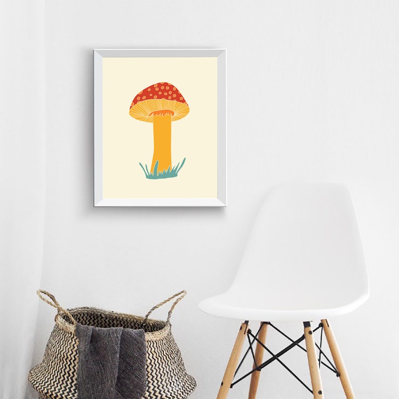 May include: A framed art print showing a stylised mushroom illustration. The mushroom has a red cap with white spots and a yellow stem, set against a cream background. Green grass is at the base of the mushroom.