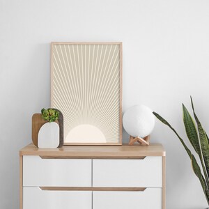 Puede incluir: Una impresi&oacute;n enmarcada de un dise&ntilde;o de sol beige y blanco en una pared blanca. La impresi&oacute;n est&aacute; colgada sobre una c&oacute;moda blanca con una tapa de madera. Una peque&ntilde;a planta en maceta y una l&aacute;mpara de luna blanca est&aacute;n sobre la c&oacute;moda.