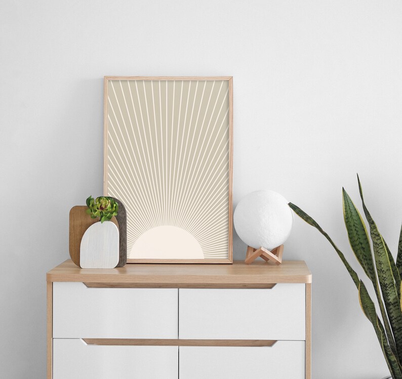 Puede incluir: Una impresi&oacute;n enmarcada de un dise&ntilde;o de sol beige y blanco en una pared blanca. La impresi&oacute;n est&aacute; colgada sobre una c&oacute;moda blanca con una tapa de madera. Una peque&ntilde;a planta en maceta y una l&aacute;mpara de luna blanca est&aacute;n sobre la c&oacute;moda.