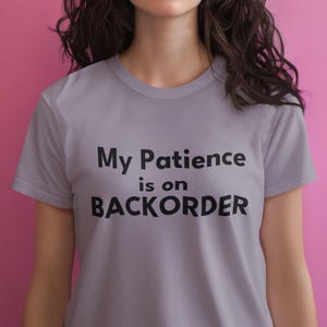 Könnte beinhalten: Hellgraues T-Shirt mit dem schwarzen Text "My Patience is on BACKORDER". Das Shirt hat kurze Ärmel und einen Rundhalsausschnitt. Der Hintergrund ist einfarbig rosa.