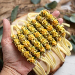 May include: Several yellow floral headbands with small yellow roses and greenery are displayed. The headbands are on a wooden surface, with a hand holding them. The background includes greenery and a burlap texture.