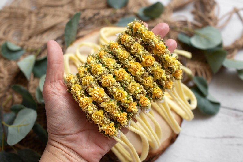 May include: Several yellow floral headbands with small yellow roses and greenery are displayed. The headbands are on a wooden surface, with a hand holding them. The background includes greenery and a burlap texture.