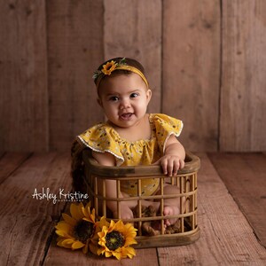 Op de afbeelding: Een baby in een gele bloemenromper en bijpassende haarband met zonnebloemdetail, zittend in een klein houten mandje. Twee zonnebloemen staan linksonder. De achtergrond is een houten muur.