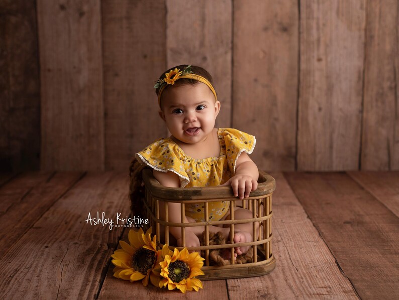 Op de afbeelding: Een baby in een gele bloemenromper en bijpassende haarband met zonnebloemdetail, zittend in een klein houten mandje. Twee zonnebloemen staan linksonder. De achtergrond is een houten muur.