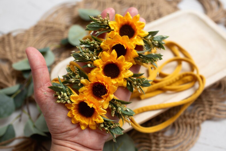 Op de afbeelding: Een mosterdgele zonnebloem haarband met vier felgele zonnebloemen en groene bladeren. De haarband is gemaakt van een zachte, mosterdkleurige stof. De bloemen hebben donkerbruine centra.